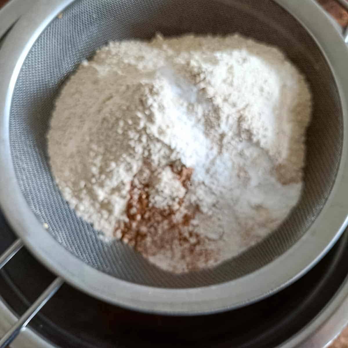 Sifted flour, baking powder, baking soda, cinnamon, and salt in a mesh sieve for vegan banana pecan muffins