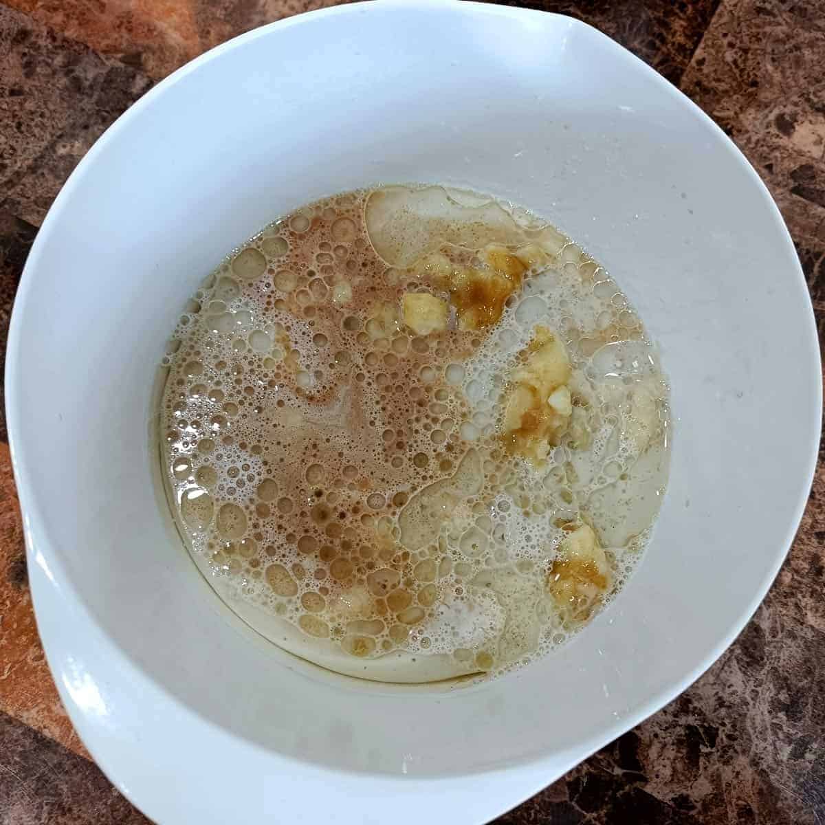 Almond milk, oil, vanilla, vinegar, sugar, and mashed banana added to a white bowl for vegan banana pecan muffins