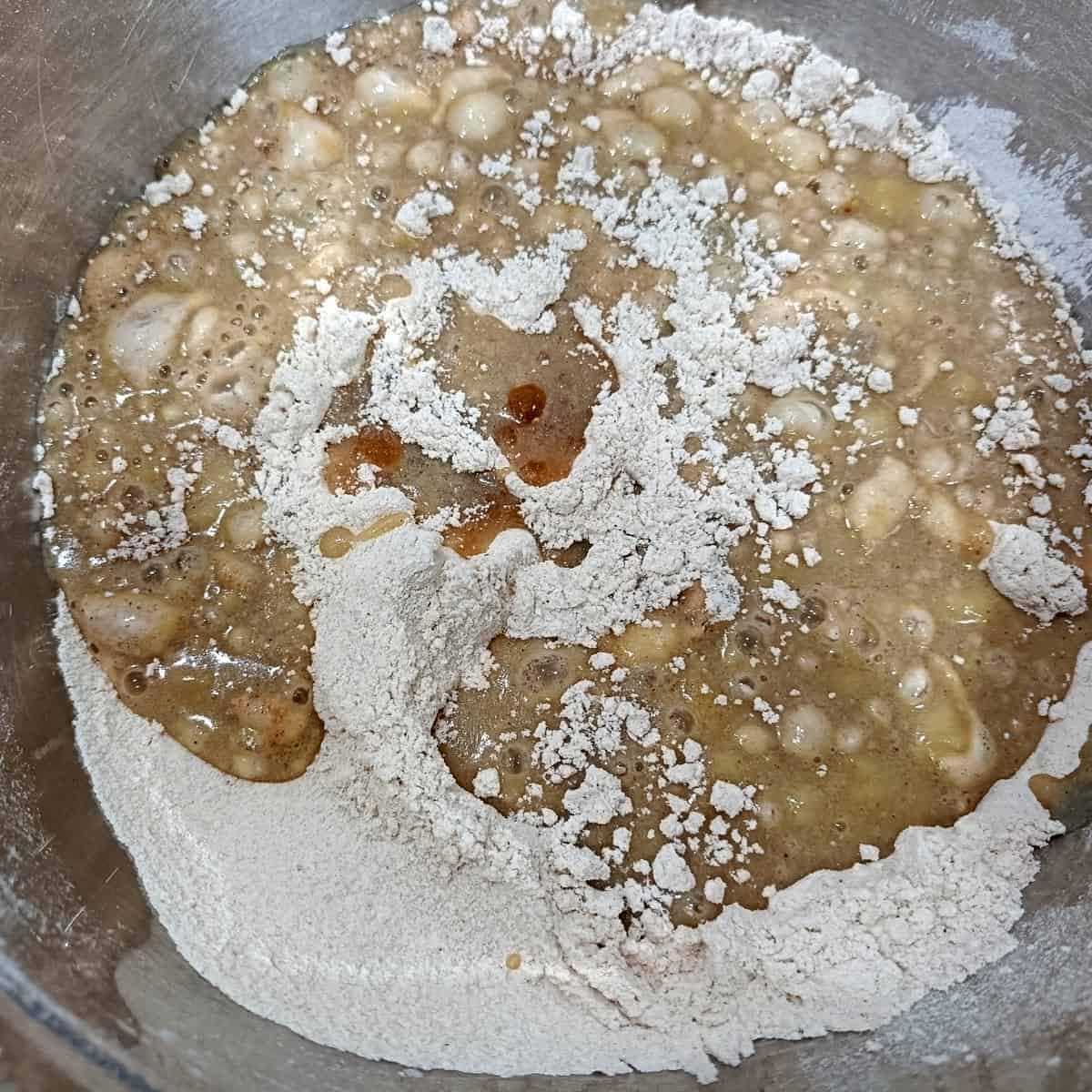 Wet banana mixture poured into sifted dry ingredients in a mixing bowl for vegan banana pecan muffins
