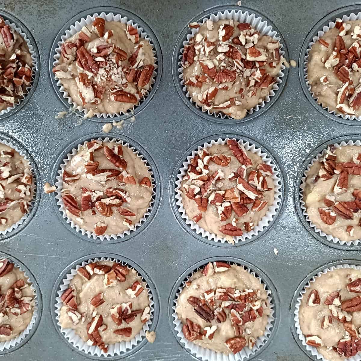 Unbaked vegan banana muffins topped with chopped pecans in a paper-lined muffin pan