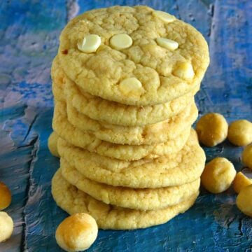 A neat stack of thick, chewy eggless white chocolate macadamia cookies highlights their tender crumb, pale golden color, and generous pockets of white chocolate, with whole macadamias scattered alongside for a rustic finish.