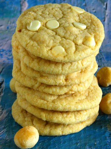 A neat stack of thick, chewy eggless white chocolate macadamia cookies highlights their tender crumb, pale golden color, and generous pockets of white chocolate, with whole macadamias scattered alongside for a rustic finish.