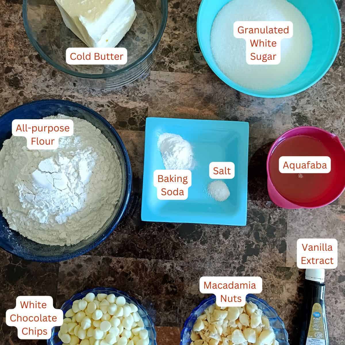 A neatly arranged overhead shot displays the measured ingredients for eggless white chocolate macadamia cookies—cold butter, sugar, flour, aquafaba, baking soda, salt, vanilla extract, white chocolate chips, and chopped macadamia nuts—clearly labeled and ready for baking.