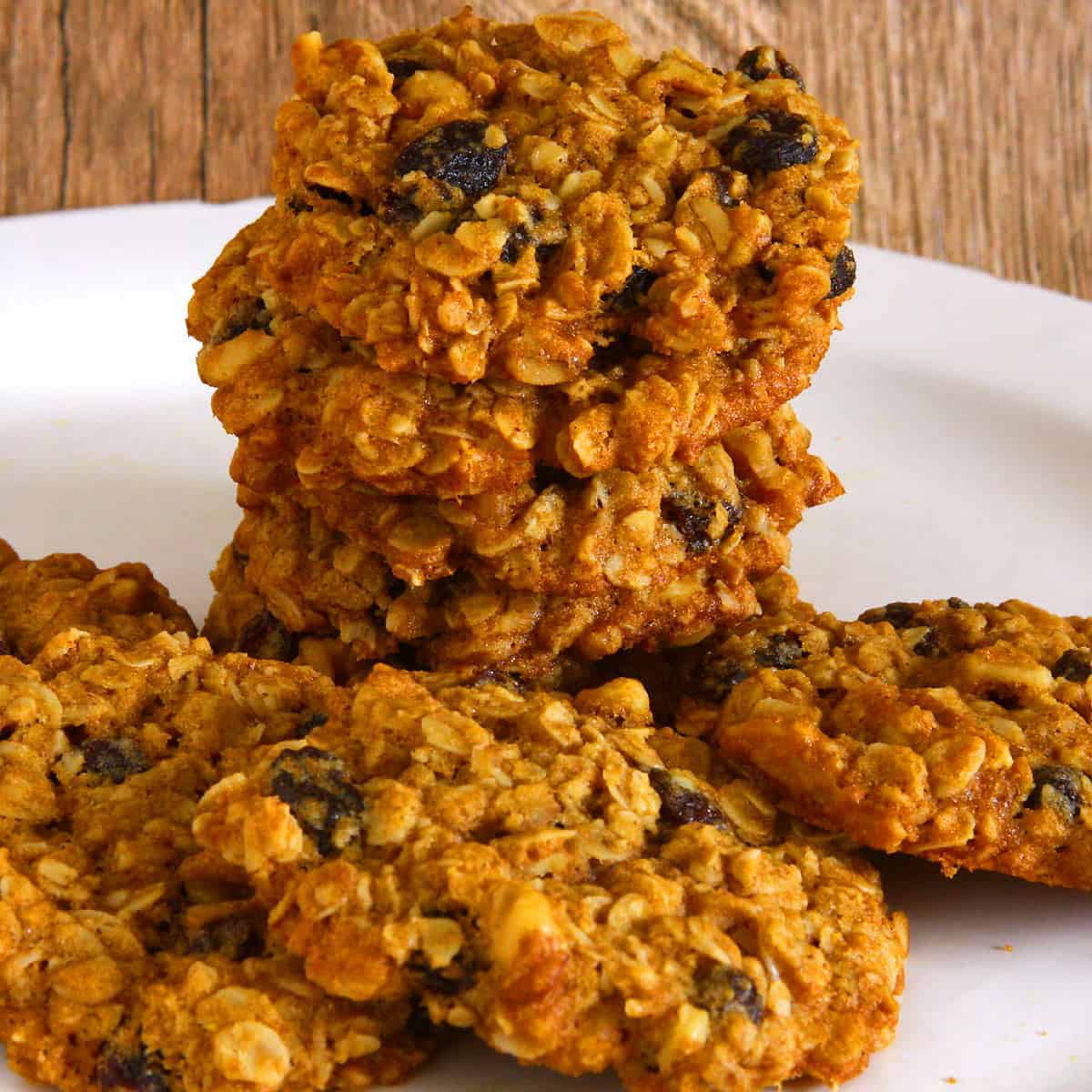 A close-up of stacked vegan oatmeal raisin cookies, golden brown with visible oats and plump raisins, showing a crisp exterior and hearty, chewy texture on a white plate.