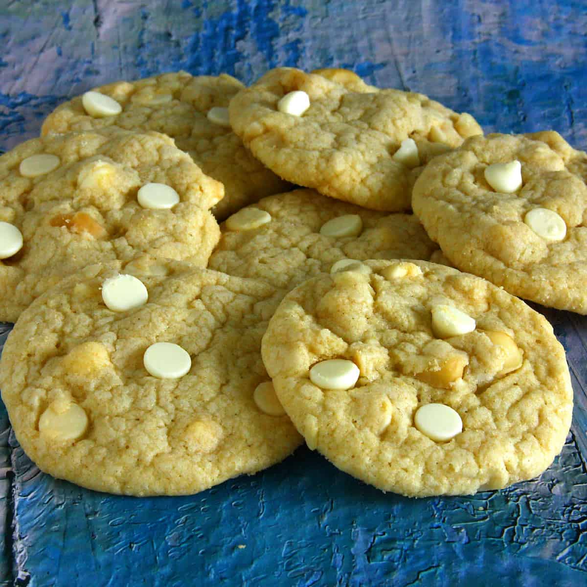 Soft, golden eggless white chocolate macadamia cookies are spread out casually, showing their slightly crinkled tops studded with creamy white chocolate chips and nutty macadamia pieces.