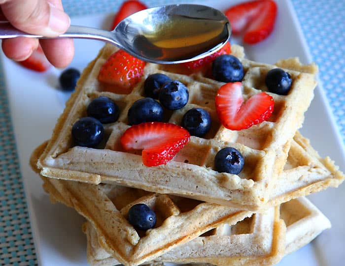 Stack of eggless waffles with fresh strawberries and blueberries, shown in soft natural light as maple syrup is drizzled from a spoon.